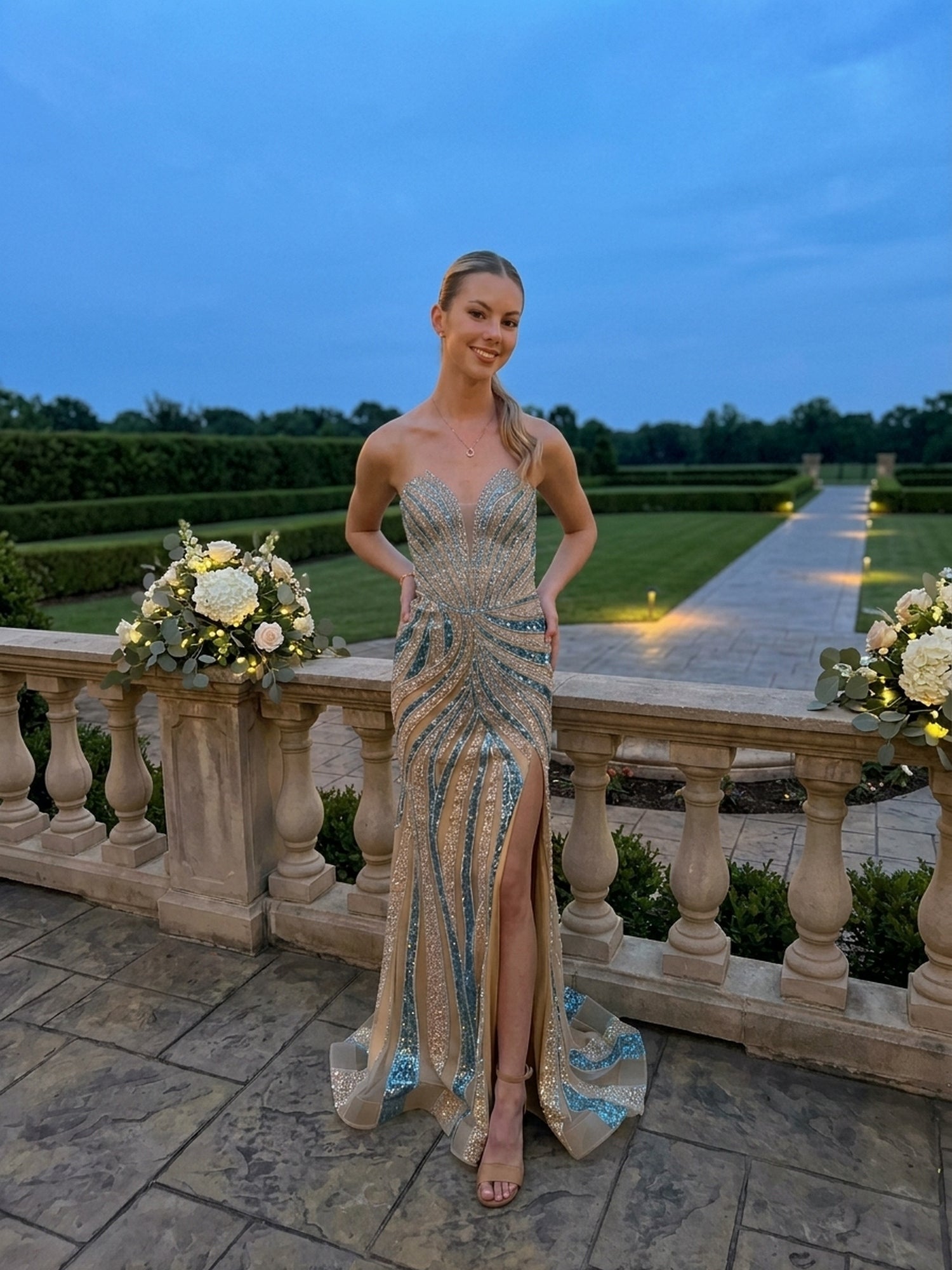 A person wearing the Long Prom Dress: Amelia Couture 2167, a retro glam, strapless, champagne and teal sequined floor-length gown with a thigh-high slit, stands on a stone terrace.